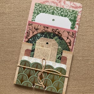 May include: A handmade paper craft project featuring a wooden card holder with various decorative papers. The papers include floral and geometric patterns in shades of green, pink, and brown, tied with twine. The project is displayed on a textured, tan surface.