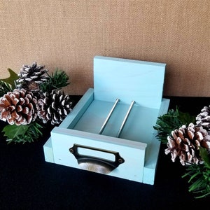 May include: A light blue wooden napkin holder with a drawer and two metal rods. The drawer has a brass pull.