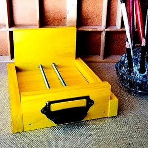 May include: A bright yellow wooden desk organizer with two silver metal rods and a black handle. A clear glass bowl filled with pencils is to the right, and a wooden shelf unit is in the background.