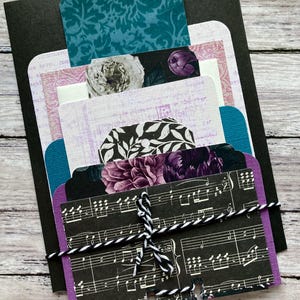 May include: A layered paper craft project featuring various patterned cards and tags in shades of teal, purple, and black. The cards are tied together with black and white twine. Floral and musical note designs are visible.
