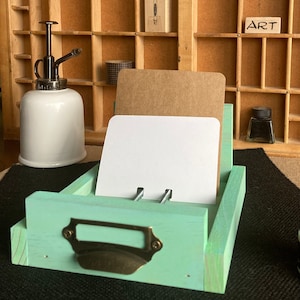 May include: A mint green wooden card holder with a metal label holder and a bronze-colored handle. The holder contains a white and a brown card. A white and bronze plant mister is to the left.