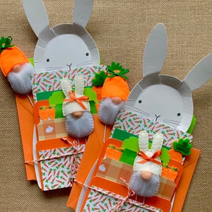 May include: Two white bunny-shaped paper plates with orange carrot and green leaf accents, each with a white bunny gnome with a red bow and a white beard, wrapped with twine and orange and white patterned paper.