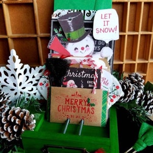 May include: A green wooden box with a snowman, pine cones, and various Christmas-themed paper crafts. The box has a metal handle and is decorated with a red bow. The paper crafts include a sign that says "Let it snow!" and a card that says "Merry Christmas to you".