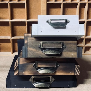May include: A stack of wooden drawers in varying shades of brown and black, each with a metal label holder and handle. The top drawer is white. The drawers are arranged in a staggered formation.