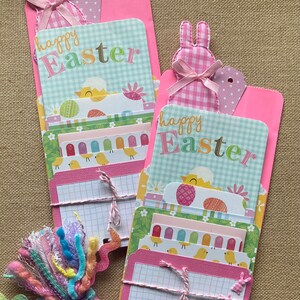 May include: Two pink folders with a bunny rabbit design and the text "happy Easter" on the front. Each folder contains a stack of colorful paper with a grid pattern and a pink ribbon tied around it.