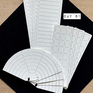 May include: A collection of white paper cards with black grid and line designs. The cards are arranged in a fan shape and rectangular stacks. The text "DAY 81" is visible on a white rectangle.