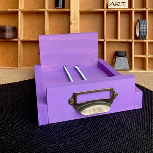 May include: Purple wooden drawer with a metal pull handle. The drawer has two slots for holding items, such as pencils or pens.