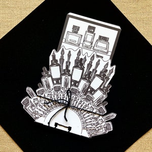 May include: Black and white illustration of a perfume bottle with a crown on top. The bottle is surrounded by smaller bottles and a variety of other objects, including a few that look like feathers.