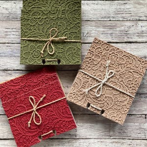 May include: Three square notebooks with lace-patterned covers in olive green, red, and beige. Each notebook is tied with twine and a bow. The notebooks are stacked on a light-colored wooden surface.