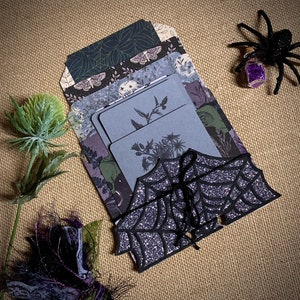 May include: A collection of decorative cards and tags with a Halloween theme. The cards feature botanical illustrations and a bat-shaped tag with a glittery web design. A black spider with a purple gem is also present, along with dried flowers, all on a tan background.