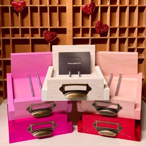 May include: Four wooden drawer boxes with metal pulls. The top box is white with a black card that says "Catalogdex Co.". The other boxes are pink, red, and magenta. The boxes are stacked on top of each other.