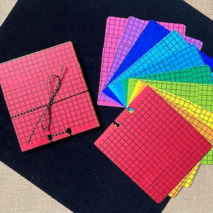 May include: A set of ten colorful gridded paper squares, arranged in a fan shape. The squares are red, orange, yellow, green, blue, indigo, and violet. The squares are tied together with a black string.