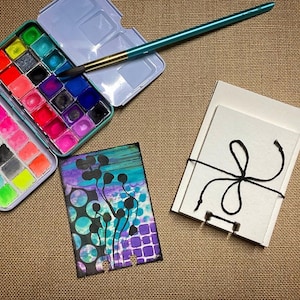 May include: A watercolor paint set with a variety of colors in a metal case, a paintbrush, a blank card with a black ribbon tied around it, and a watercolor painting of abstract shapes in shades of blue, purple, and green.