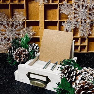 May include: A white wooden drawer with a brass handle, used as a card holder. The drawer is filled with brown cards and sits on a black surface. The drawer is decorated with pine branches and pine cones.
