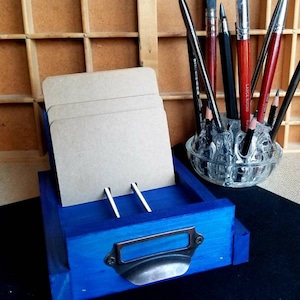 May include: A blue wooden card holder with a metal drawer pull. The holder has three slots for cards and is sitting on a black surface. There are several paintbrushes and pencils in a glass jar behind the holder.