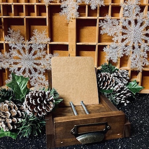May include: A wooden box with a drawer, decorated with a vintage style metal handle. The box is filled with brown card stock paper. The box is surrounded by white snowflake ornaments and pine branches.