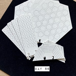 May include: A set of white paper templates with different geometric patterns, including hexagons, squares, and triangles. The templates are arranged in a fan shape on a black surface. The text "DAY 86" is printed on the surface below the templates.