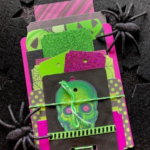 May include: A Halloween-themed craft project featuring a stack of decorative tags and cards in shades of green, pink, and black. The focal point is a green skull illustration. Two glittery black spiders flank the stack, adding to the spooky aesthetic.