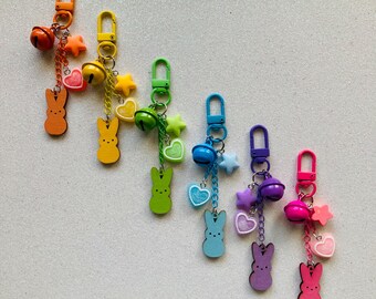 Peut inclure: Un ensemble de sept porte-clés colorés avec un charme en forme de lapin, des cloches et des charmes en forme d'étoile. Les porte-clés sont disposés en arc-en-ciel, chaque porte-clés ayant une couleur différente.