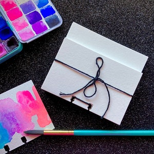 May include: A small, white, blank notebook with a black string tied around it. The notebook is sitting on a black surface with a watercolor paint set and a paintbrush.