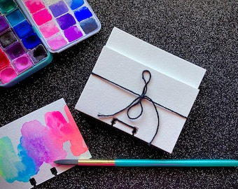 May include: A small, white, blank notebook with a black string tied around it. The notebook is sitting on a black surface with a watercolor paint set and a paintbrush.