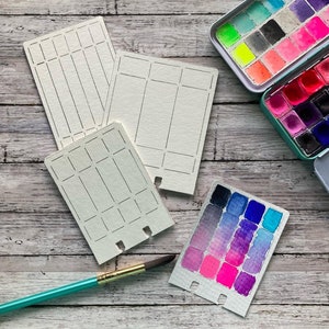 May include: Three white watercolor paper cards with grid lines and a paint swatch card with a gradient of pink, purple, and blue watercolor paint. The cards have perforated edges and are on a wooden surface.