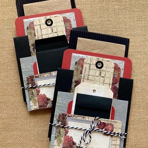 May include: Two handmade paper card sets. Each set includes a red, black, and tan card, a tag, and a small black envelope. The cards are decorated with floral and vintage-style paper. The sets are tied with black and white twine.