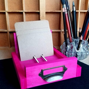 May include: A bright pink wooden card holder with a drawer and a metal handle. The holder has three slots for cards and is sitting on a black surface.