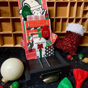 May include: A black wooden drawer with a metal handle, filled with colorful Christmas cards and decorations. The drawer is sitting on a black surface with red and green glitter ornaments.