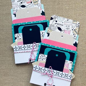 May include: Two handmade paper card holders with coffee-themed designs. Each holder has multiple layers of cards in various colors, including pink, teal, and black. The top layer features a coffee cup pattern, and the holders are tied with red and white twine.