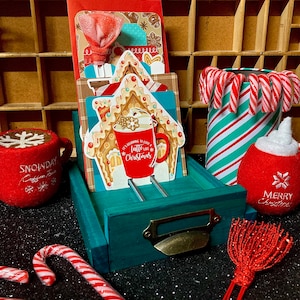 May include: A teal blue wooden drawer with a brass handle, holding a stack of Christmas cards featuring a gingerbread house and a red coffee mug with the text "It's beginning to look a latte like Christmas". The drawer is surrounded by red and white candy canes, a red and white coffee mug with a snowflake design, and a red felt ornament with the text "Merry Christmas".