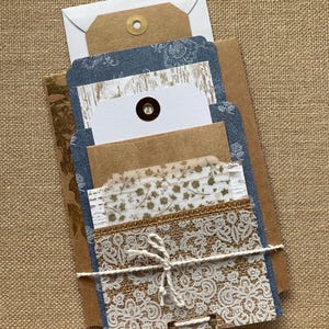 May include: A collection of handmade stationery items, including envelopes, tags, and decorative cards. The cards feature a mix of textures, including lace, burlap, and patterned paper, tied together with twine. The color palette is neutral with touches of blue.
