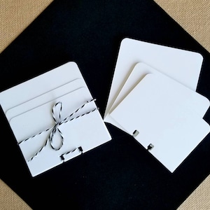 May include: A set of three white cardstock notecards with a black and white twine bow. The notecards are folded in half and have a small hole punched in the top corner.