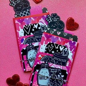 May include: A stack of handmade Valentine's Day cards with a pink and black color scheme. The cards feature cute illustrations of ice cream cones, milk bottles, coffee cups, and donuts. The cards are tied together with a white ribbon and decorated with red hearts.