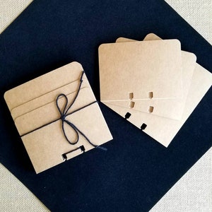 May include: A set of four brown kraft paper cards with rounded corners and three holes punched along the top edge. The cards are tied together with black twine and are arranged on a black surface.