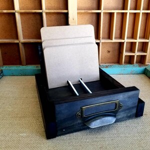 May include: A dark wooden drawer with a metal handle and a label. The drawer is open and contains several pieces of cardboard.