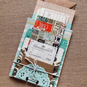 May include: A handmade paper craft project featuring a collection of vintage-style ephemera. The design includes license plates, receipts, and a memorandum card, all tied together with twine and a light blue paper doily. A small brown envelope is also included.