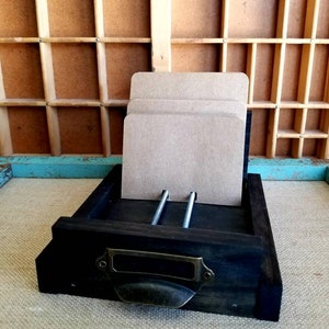 May include: A dark wooden drawer with a metal handle and two metal screws. The drawer is holding a stack of brown paper cards.