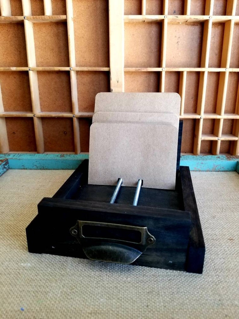 May include: A dark wooden drawer with a metal handle and two metal screws. The drawer is holding a stack of brown paper cards.