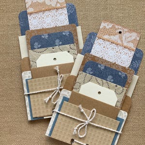 May include: Two handmade paper pocket sets. Each set includes various patterned cards, tags, and decorative elements. The pockets are tied with twine. The sets feature a mix of neutral tones, blue floral patterns, and lace accents, all arranged on a textured beige surface.