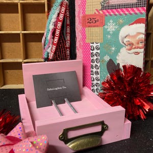 May include: A pink wooden card holder with a brass handle and a black card that reads "Catalogdex Co." is in front of a Christmas-themed journal. The journal has a Santa Claus image and a "25¢" ticket.