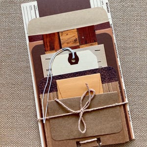 May include: A collection of brown and tan paper ephemera, including tags, envelopes, and patterned paper, tied together with twine. The items are arranged in a layered fashion, with a dark brown tag at the top.