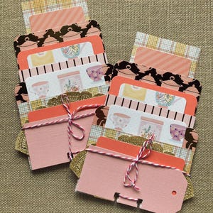 May include: Two stacks of pink and patterned cards tied with pink and white twine. The cards feature illustrations of teacups, teapots, and floral designs. The background is a neutral tan fabric.