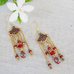 Può includere: Un paio di orecchini pendenti color oro con intricate perline rosse e verdi. Ogni orecchino presenta un elemento geometrico centrale con catenelle dorate, perline di cristallo rosso sfaccettato e perline di vetro rosse e verdi.