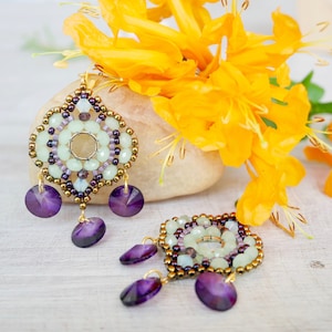 Può includere: Un paio di orecchini di perline con un design circolare centrale composto da perline verde chiaro, circondato da perline color bronzo metallizzato e viola scuro. Ogni orecchino include tre cristalli viola sfaccettati pendenti.