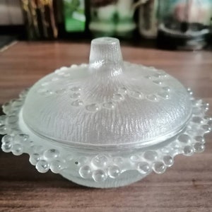 Pode incluir: Um frasco de vidro transparente com acabamento fosco e uma tampa abobadada. O frasco tem uma borda com contas e uma superfície texturizada.