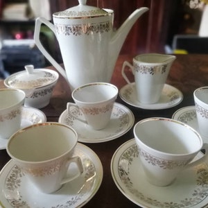 Może przedstawiać: Biały porcelanowy zestaw do herbaty z pozłacanymi zdobieniami. Zestaw zawiera czajnik, dzbanek do śmietanki, cukiernicę i cztery filiżanki z talerzykami.
