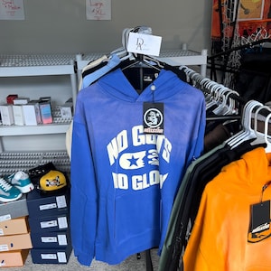 Pode incluir: Um moletom azul com o texto "NO GUYS IO GLOY" em branco. O moletom tem um bolso frontal e um capuz. Uma etiqueta preta com a marca "HELLSTAR" está anexada. Outros moletons em várias cores são visíveis.