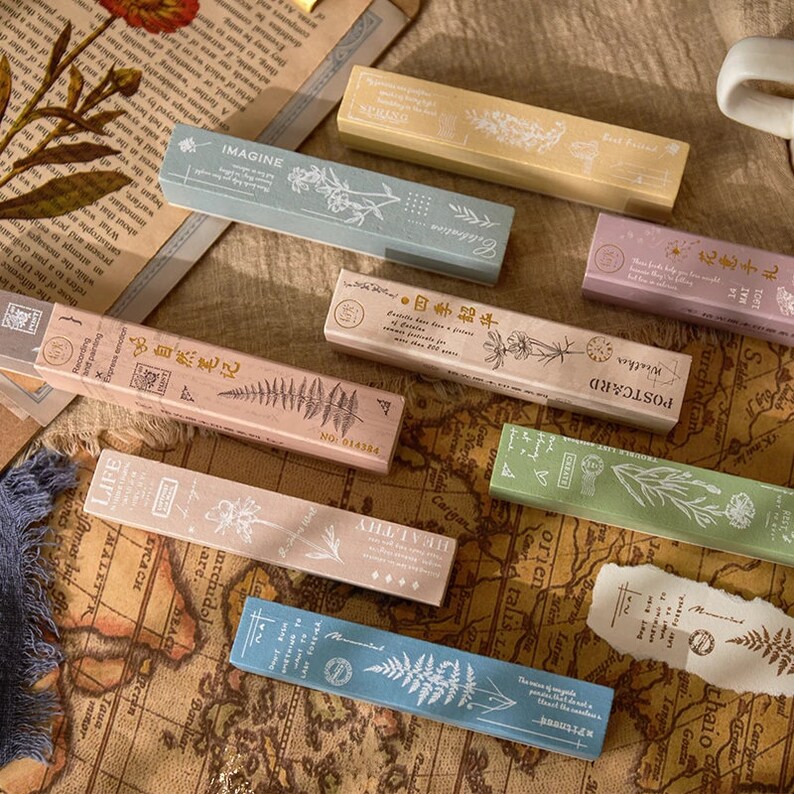May include: A set of ten wooden stamps featuring floral and botanical designs. The stamps are rectangular and have a light brown, pink, blue, and green colour scheme. The stamps feature text such as "Imagine", "Celebration", "Postcard", "Weather", and "Life".