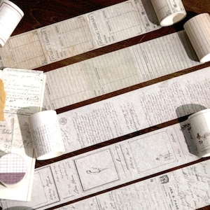 May include: Assortment of decorative washi tapes with various patterns and designs. The tapes are white with black and gray text and illustrations. The tapes are rolled up and some are partially unrolled, revealing the designs.